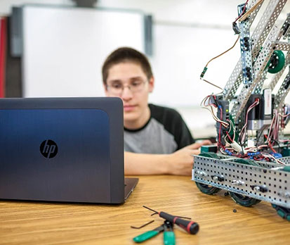 Student using a laptop to work on a metal robotics build with tools on the table.