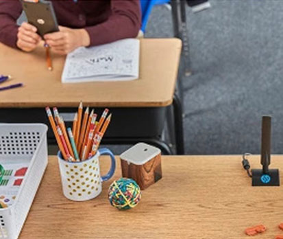 Close-up of a student desk with pencils in a cup, a rubber band ball, and school supplies while a student works in the background.