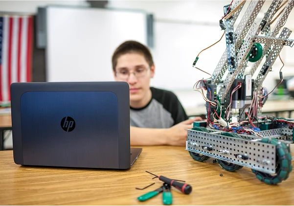 Student working on a robot with a laptop and tools in a classroom setting.