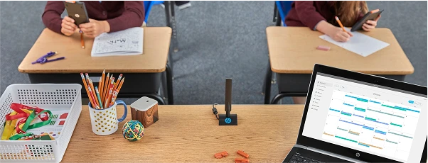 Classroom desk with supplies, a laptop displaying a calendar, and students in the background working with pencils and smartphones.
