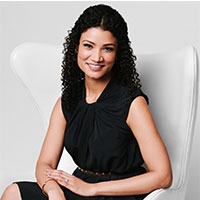 Smiling woman with curly hair wearing a sleeveless black dress seated on a white chair